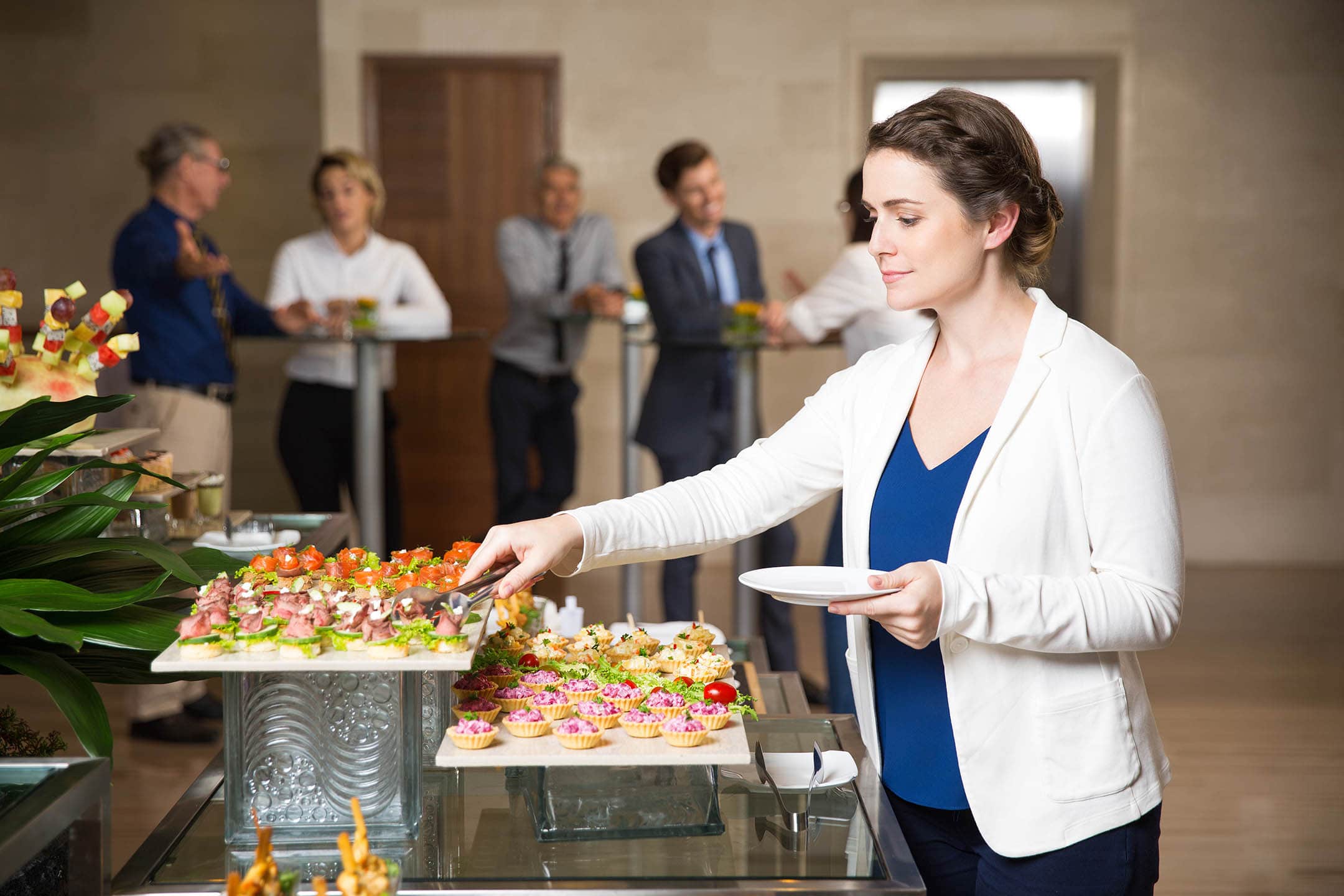 Young businesswoman serving herself with canapes. Business people eating and talking in background